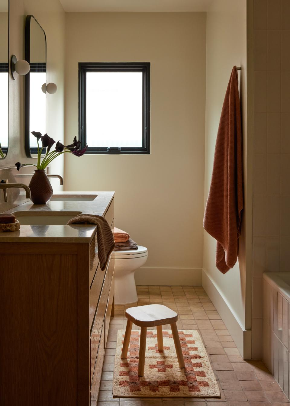 The designers updated the primary bathroom with a double vanity and sleek fixtures and finishes. Sconces by Matt Alford Studio elevate the walls. The mirrors, stool, and rug are from West Elm, Etsy, and Leif respectively. Towels are Coyuchi.
