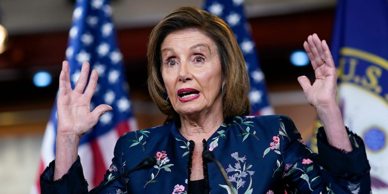 Nancy Pelosi speaking and holding up both hands at a press conference.