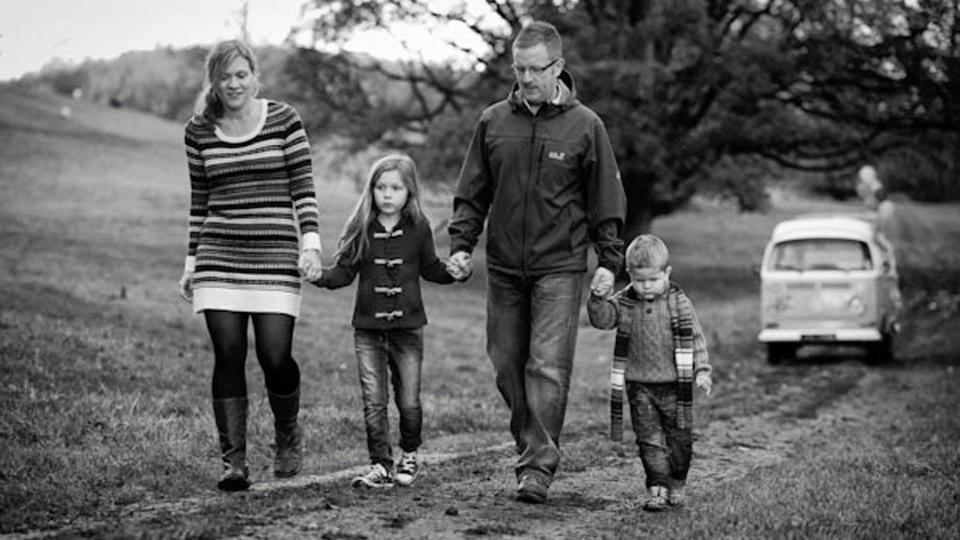 A black and white photograph of the Sherriff family in coats and scarves, holding hands as they walk along a path through a muddy field, with a VW camper van in the background.