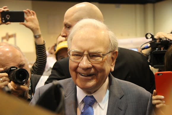 A smiling Warren Buffett in a crowd of people and reporters during a Berkshire Hathaway shareholder meeting.
