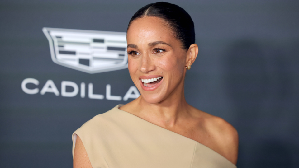 Photo of Meghan Markle smiling for photographers against a photo wall. 