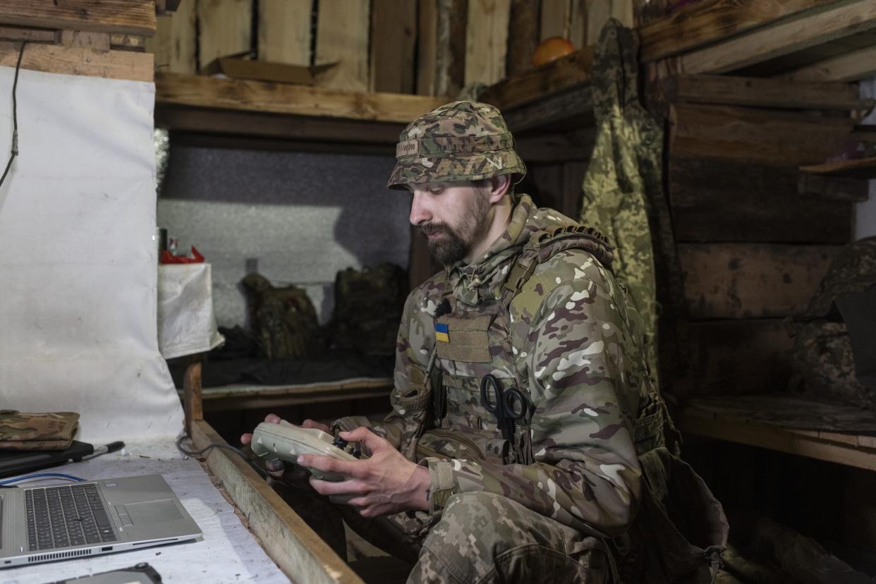 A Ukrainian soldier holding a remote controller while sitting in front of a laptop in a wooden shack.