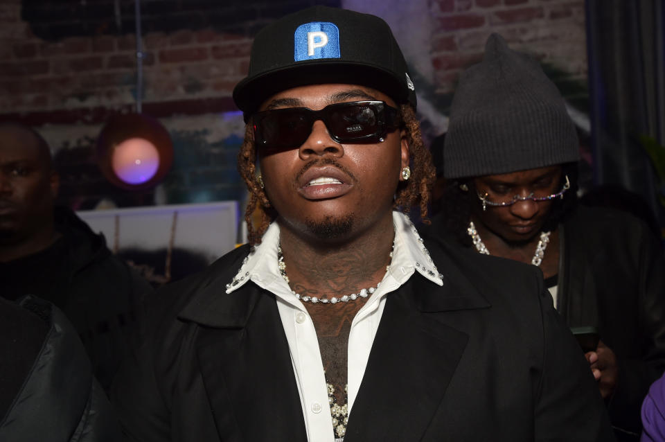 Gunna in a black jacket, white shirt, black sunglasses, and black hat with blue and white letter "P" 