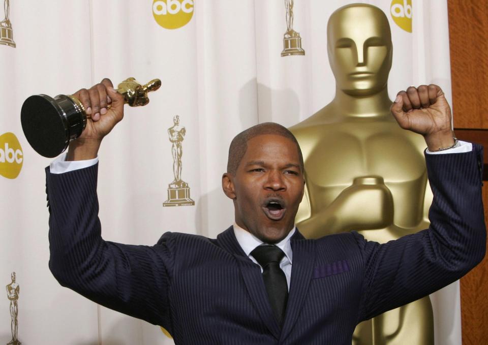 jamie foxx wearing a black suit and tie, pumping both his fists into the air, holding an oscar statuette in his right hand, and standing in front of a large oscar statue