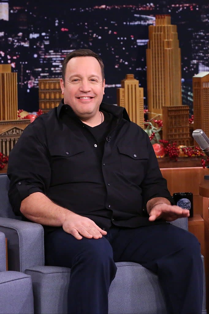 Man in a black shirt sitting on a couch, smiling, on a talk show set