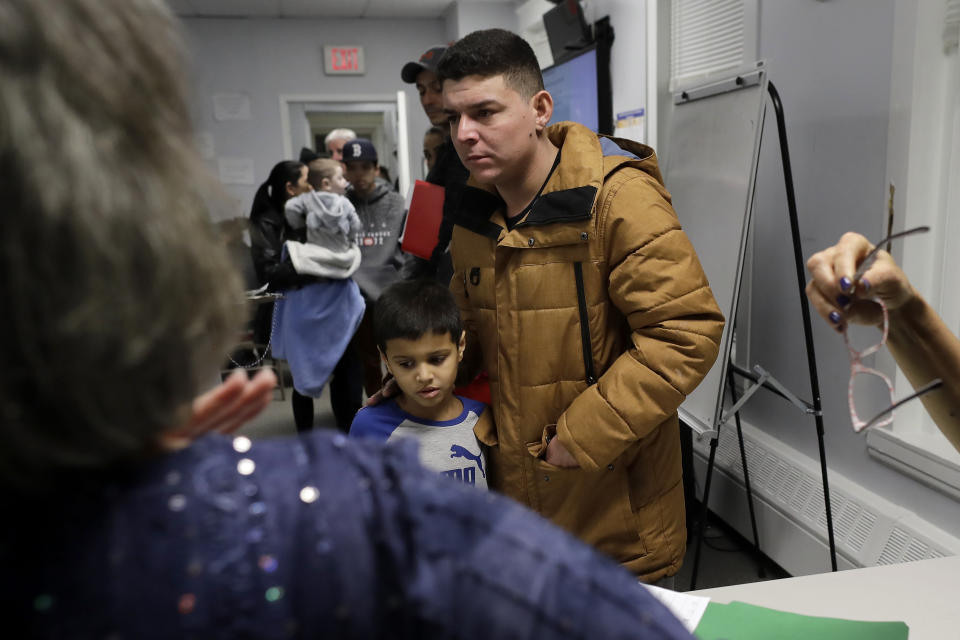 In this Wednesday, Dec. 4, 2019 photo Helison Alvarenga, of Brazil, center right, and his 6-year-old son, David, center left, move away from a reception table after speaking with volunteers at the New England Community Center, in Stoughton, Mass. Alvarenga, a 26-year-old from the Brazilian state of Minas Gerais, arrived in Massachusetts about four months ago after crossing the Mexican border at Juarez with his 24-year-old wife, Amanda, and his son David. The family were at the community center Dec. 4 to apply for new Brazilian passports, which Alvarenga says were seized by border officials. (AP Photo/Steven Senne)
