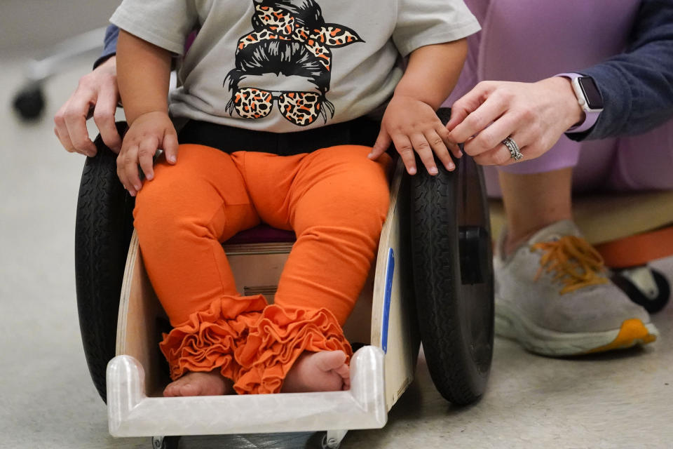 Dr. Jennifer Gaubert, a physical therapist, helps introduce Freya Baudoin,18 months, to her new mobility chair, at the Children's Hospital New Orleans Rehabilitation Center in Metairie, La., Monday, Oct. 30, 2023. Tulane science and engineering students are making the second batch of mobility chairs for toddlers, that will eventually go to pediatric patients at Children's Hospital. Wheelchairs are expensive, and insurance won't cover the cost for children unless the child proves they can operate it independently. (AP Photo/Gerald Herbert)