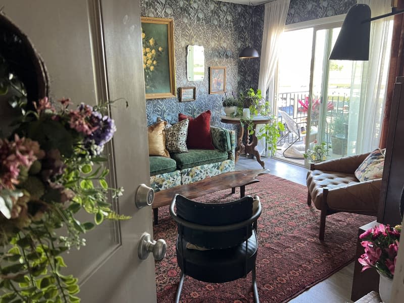 Living room with botanical wallpaper, green floral sofa and pillows, and lots of patterned textiles. Dog on the rug