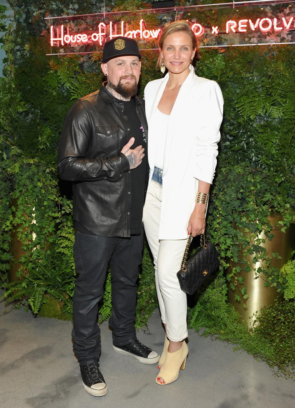 Cameron Diaz in a white blazer and pant ensemble and heels standing taller than Benji Madden in a leather jacket, hat, and Converse sneakers.