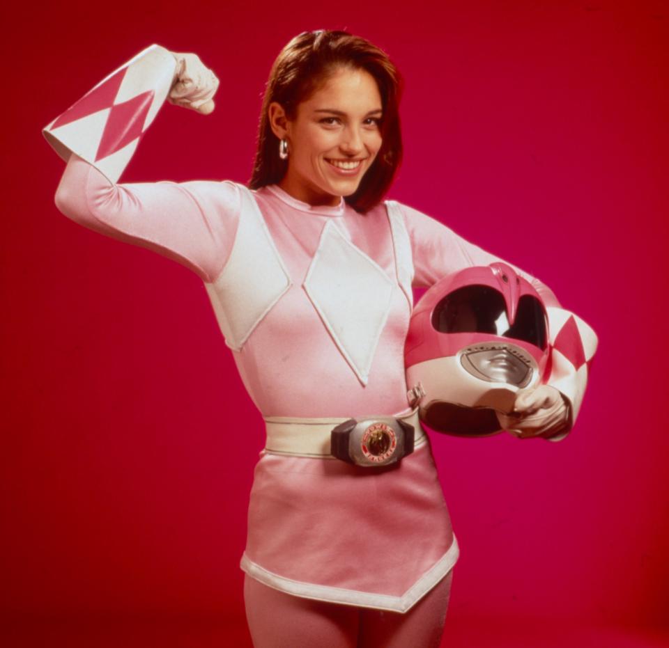 Amy Jo Johnson, dressed as the Pink Power Ranger, flexes her arm and smiles while holding the Power Ranger helmet in her other hand