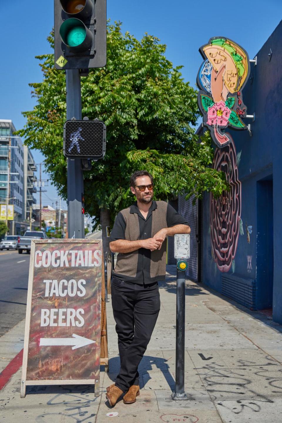 A man with dark hair, wearing sunglasses, leans on a parking meter, next to a sign advertising a restaurant's offerings.