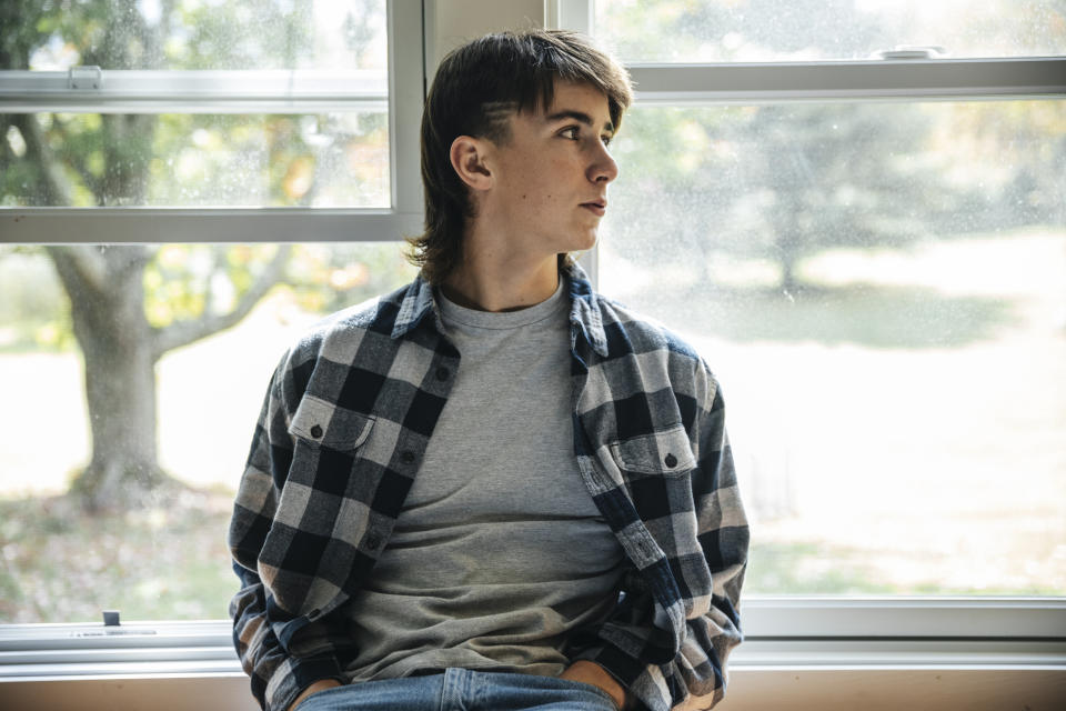 A young man wearing a plaid shirt over a t-shirt sits on a windowsill, looking out the window