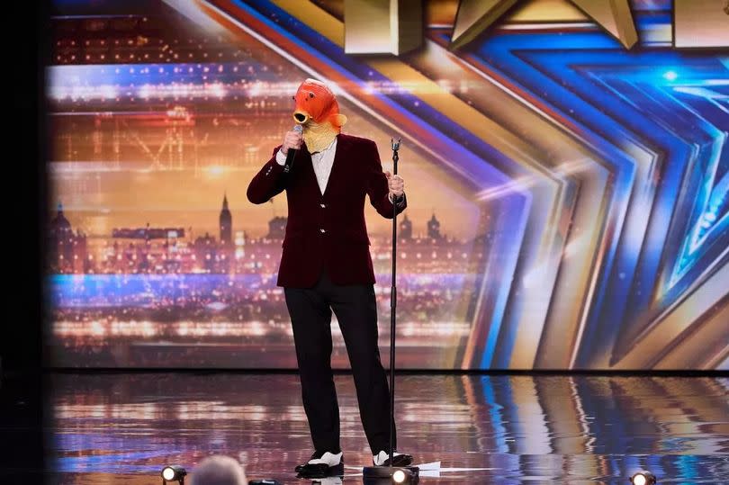 A person with an orange fish face wearing a suit and holding a microphone on Britain's Got Talent