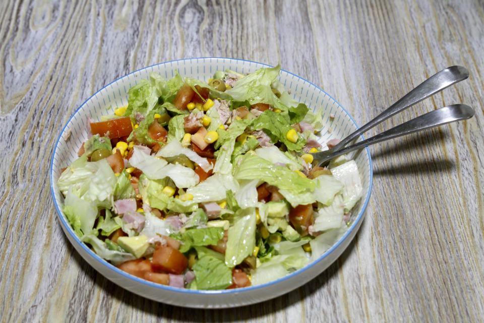 Mixed salad with lettuce, tomatoes, corn and ham cubes in a bowl, with tongs on a wooden background