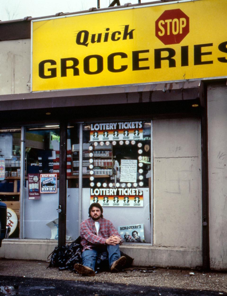 Kevin on location seated in front of a grocery store under a sign, with a reflective expression