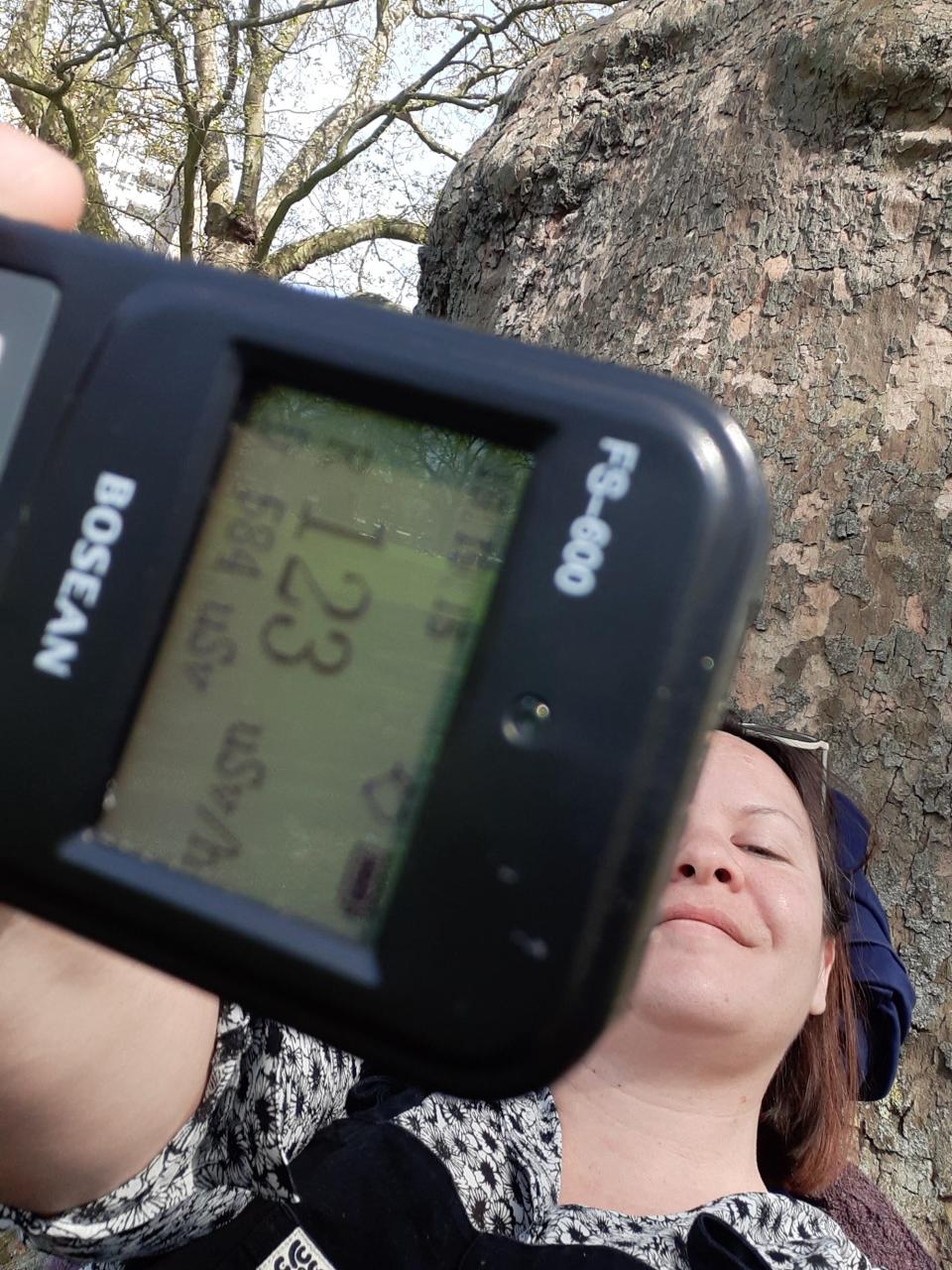 Marianne Guenot sits under a tree holding a Geiger counter at arm's length reading 123 uSv/h..