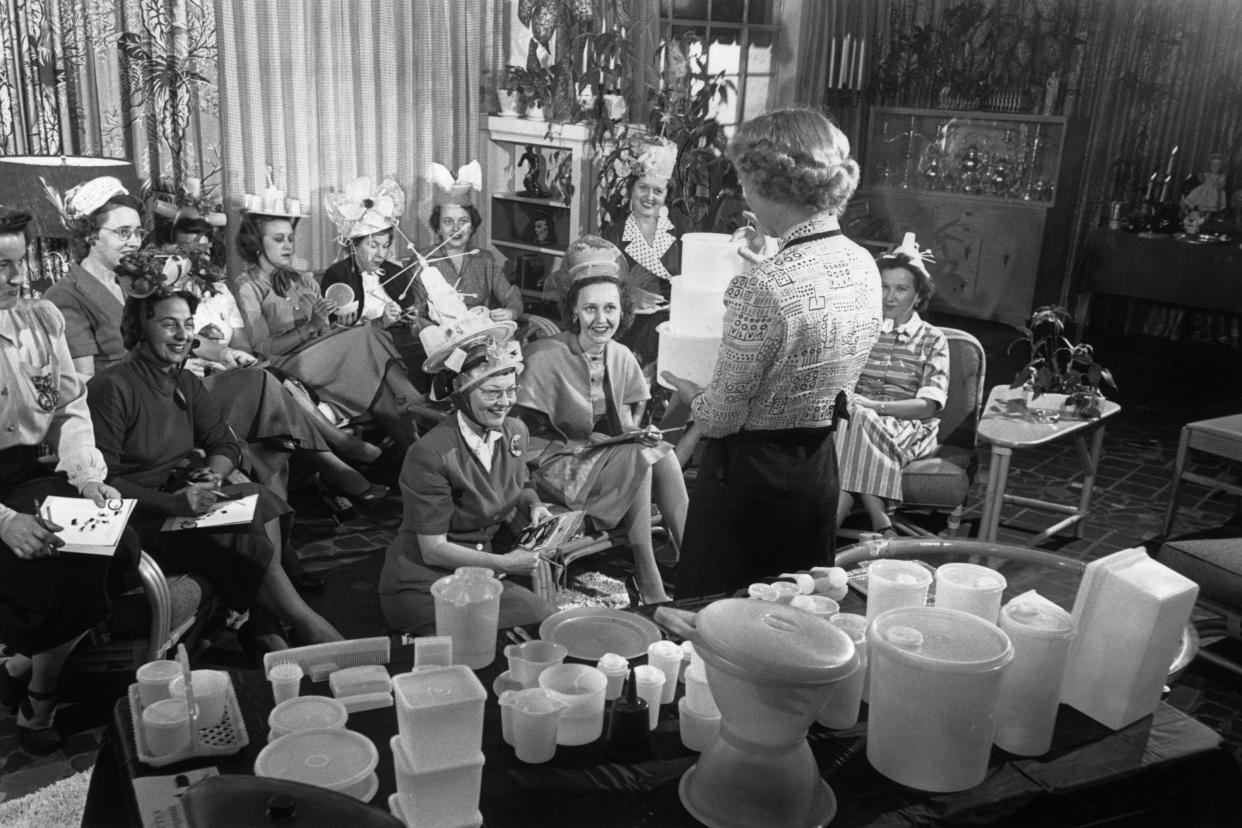 A woman holds three Tupperware containers while standing in front of a group of women seated in a living room during a Tupperware party.