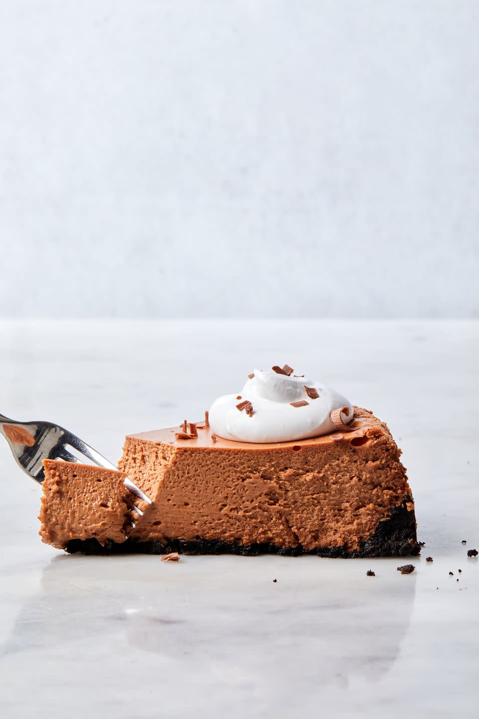 slice of chocolate cheesecake with oreo crust, topped with whipped cream and chocolate curls, with a fork taking a bite out