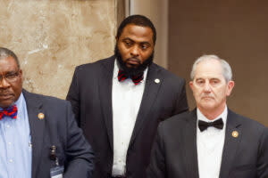 Three men in jackets and bowties stand in a marble hall.