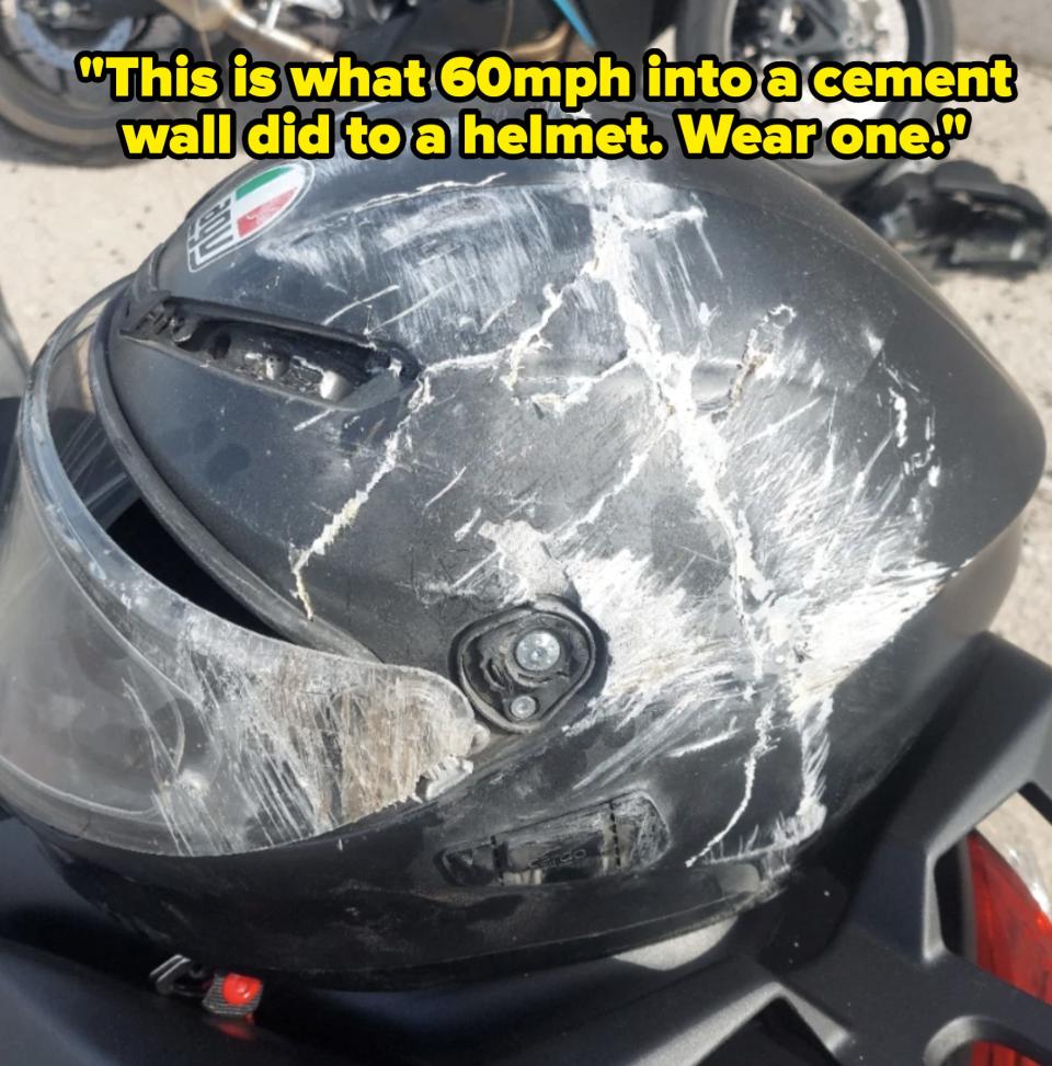 A scratched motorcycle helmet, showing significant damage. Two motorcycles are parked in the background