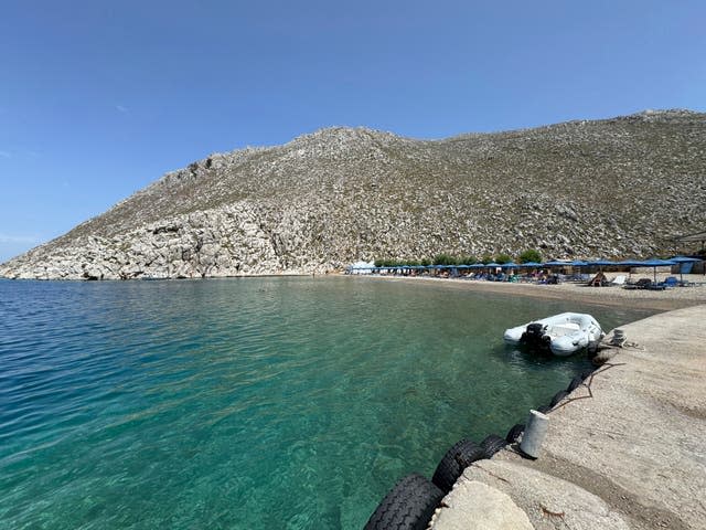 People sit on the beach of Agios Nikolaos on the Greek island of Symi with green water and a rocky backdrop