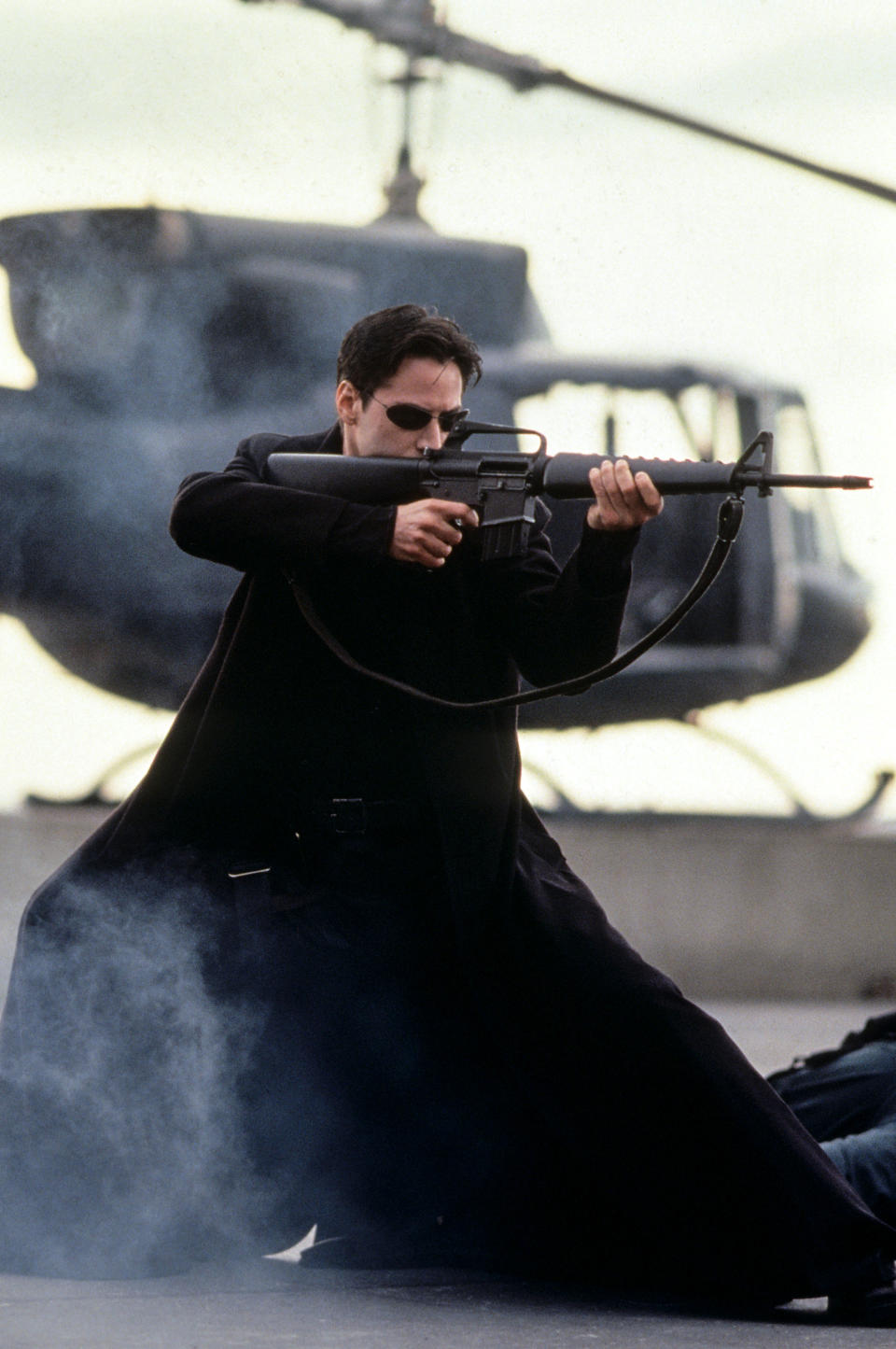 Keanu Reeves as Neo in "The Matrix" wearing a trench coat, sunglasses, and holding a rifle, with a helicopter in the background