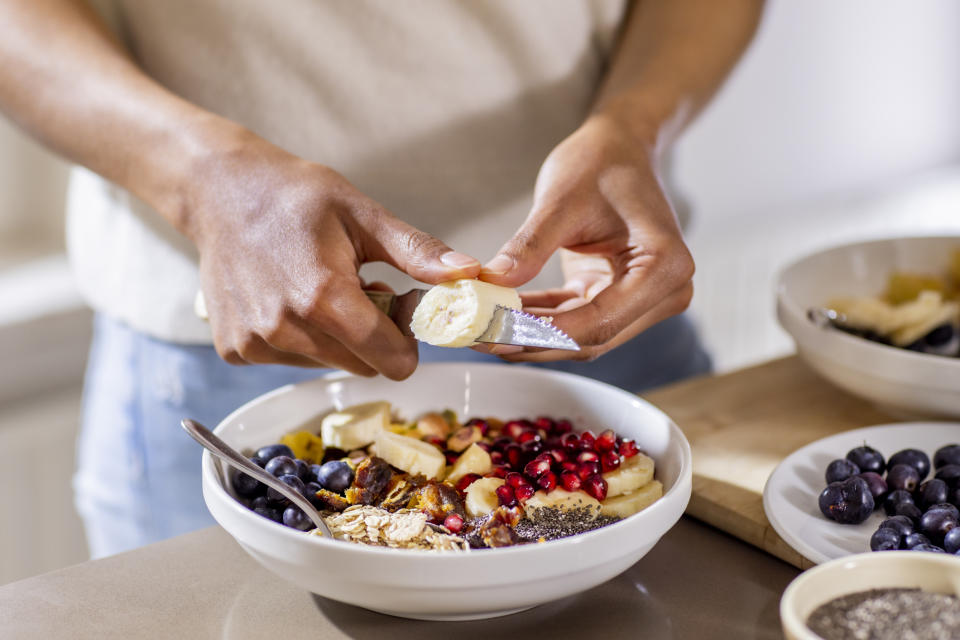 A person is slicing a banana over a bowl filled with fruit, nuts, and seeds. Other bowls with similar ingredients are on the table