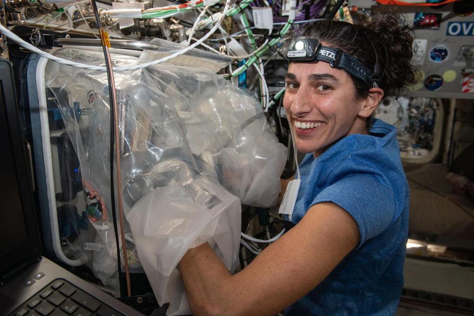 A female astronaut with a headband light on the International Space Station has her arms inside a large plastic device.