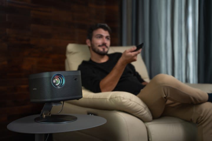 A man in a chair with a remote, and a projector on a table.