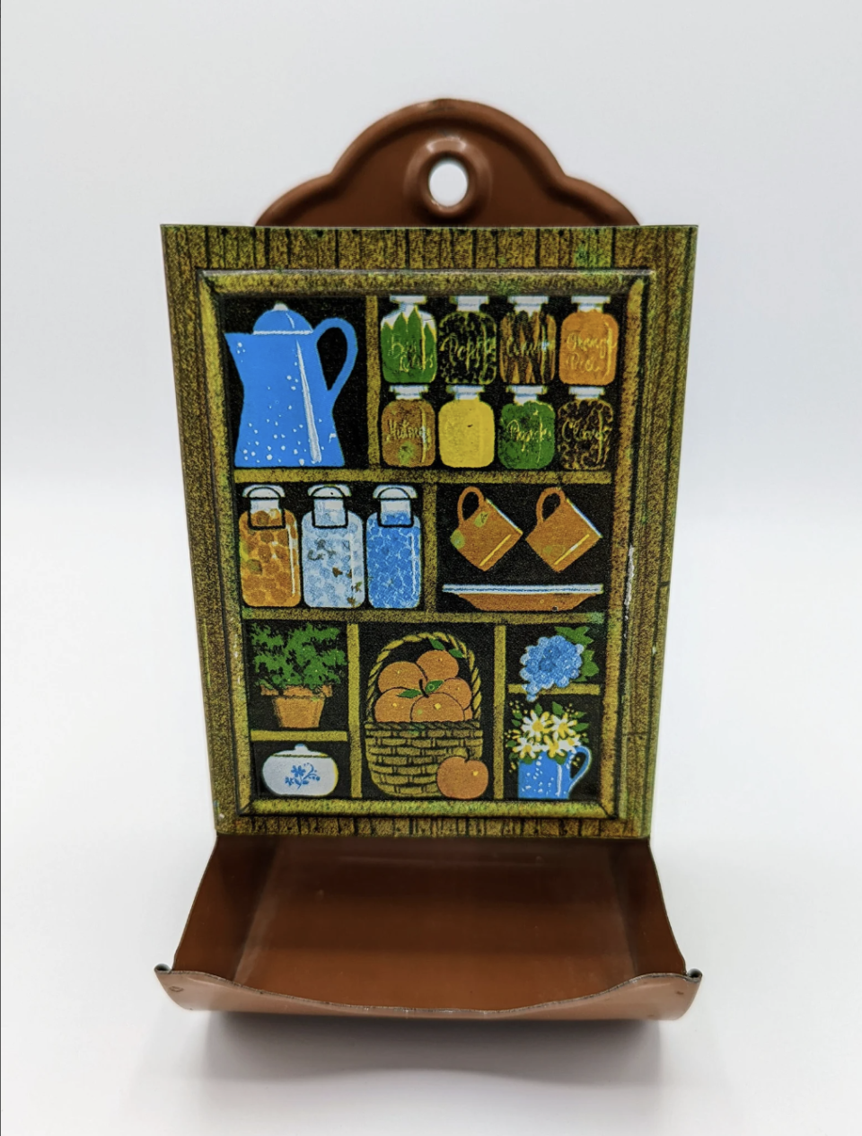 Photograph of a decorative metal wall hanging featuring painted kitchen items like jars, pitchers, and a basket of fruit on shelves