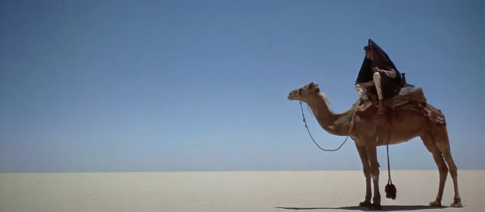 A man rides a camel across a vast, empty desert under a clear sky in a scene from a movie