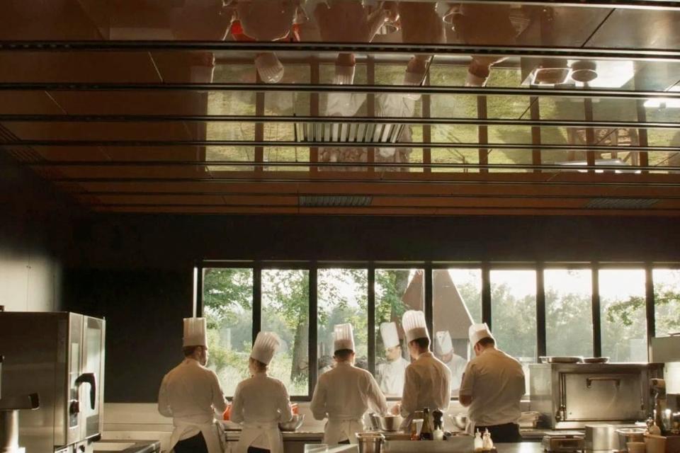 Five chefs wearing chef hats and aprons are seen from behind in a bright and high-ceilinged commercial kitchen. 