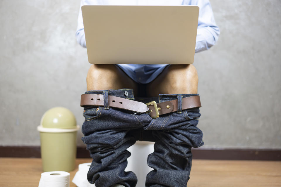 Person sitting on a toilet with jeans and belt around ankles, using a laptop on their lap. Their face is not visible