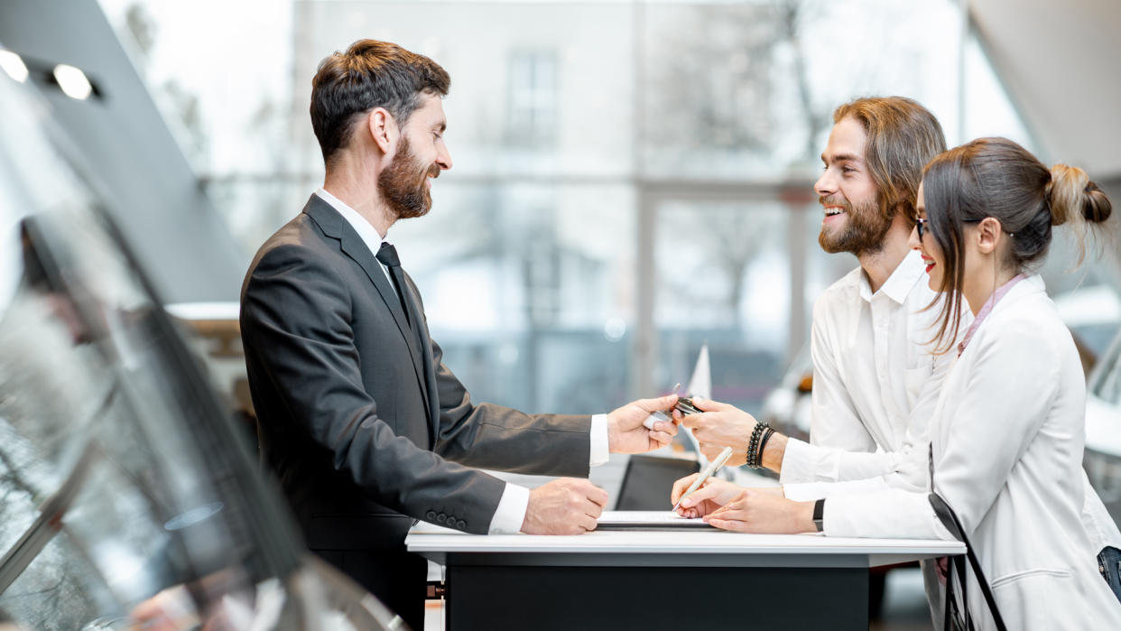 Young business couple signing some documents at the table with salesperson or manager buying or renting car in the showroom.