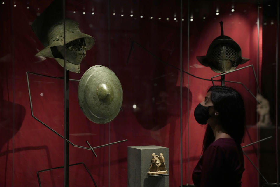 A museum employee poses for photographers next to Roman era gladiatorial helmets and a shield, during a media preview for the "Nero: the man behind the myth" exhibition, at the British Museum in London, Monday, May 24, 2021. The exhibition, which open to visitors on May 27 and runs until October 24, explores the true story of Rome's fifth emperor informed by new research and archaeological evidence from the time. (AP Photo/Matt Dunham)