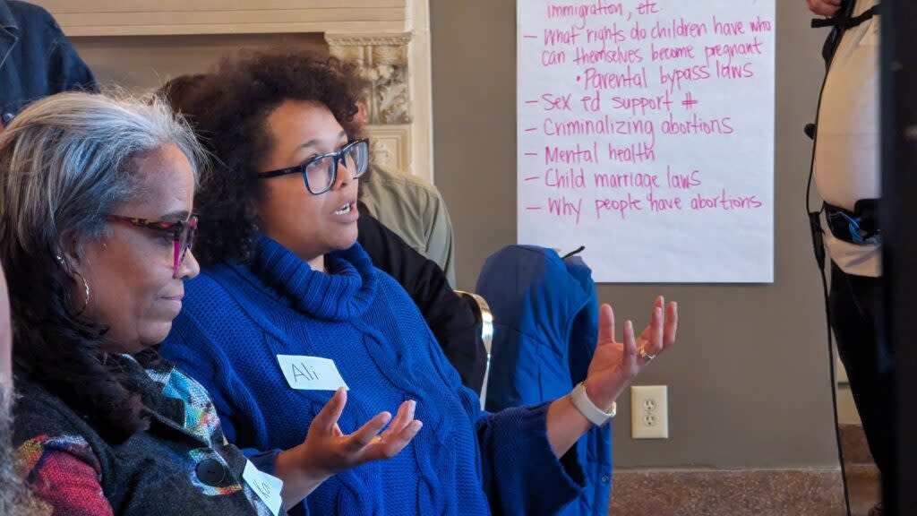 A woman in a blue sweater speaking and holding her hands up