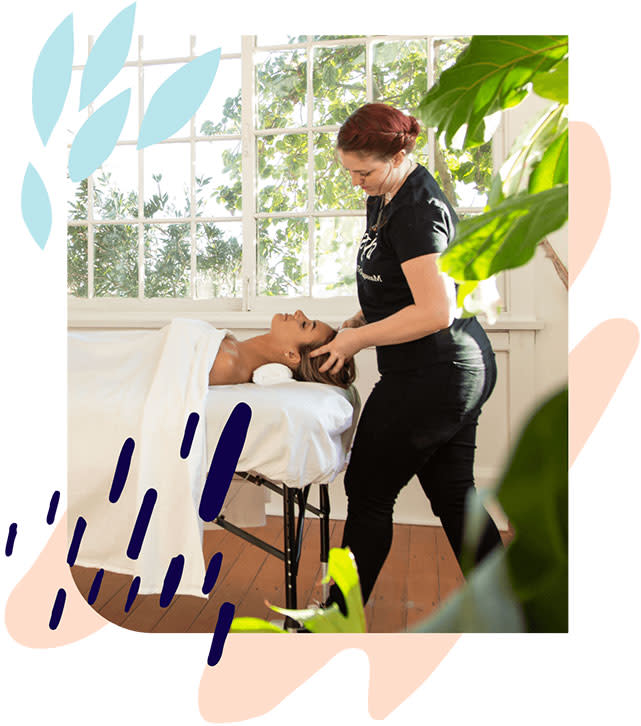A woman in black massages the head of a woman on a massage table in a light, bright room with plants and a big window.