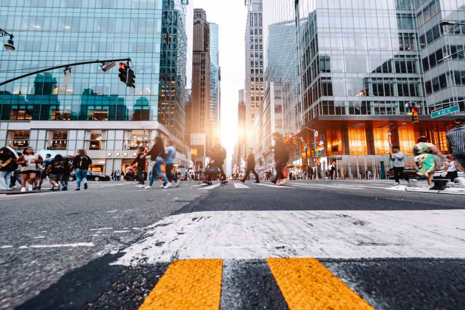 A busy city street intersection with people walking, tall buildings on either side, and sunlight in the background