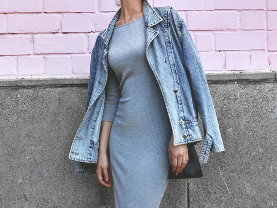 A woman in a form-fitting light blue dress with a denim jacket draped over her shoulders poses in front of a pink brick and gray wall.