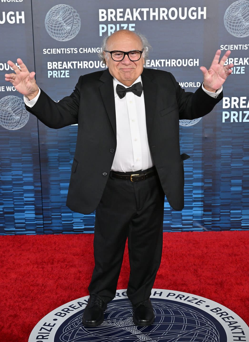 danny devito wearing a black bowtie, white shirt, a black suit jacket and trousers, and black glasses, posing on the red carpet in 2023