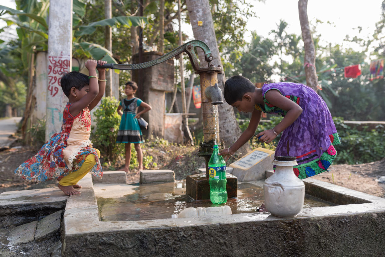 A girl in a purple dress with a green, embroidered border, leans over to fill a plastic bottle with water, while her younger sister squats on a low concrete wall to activate the pump.