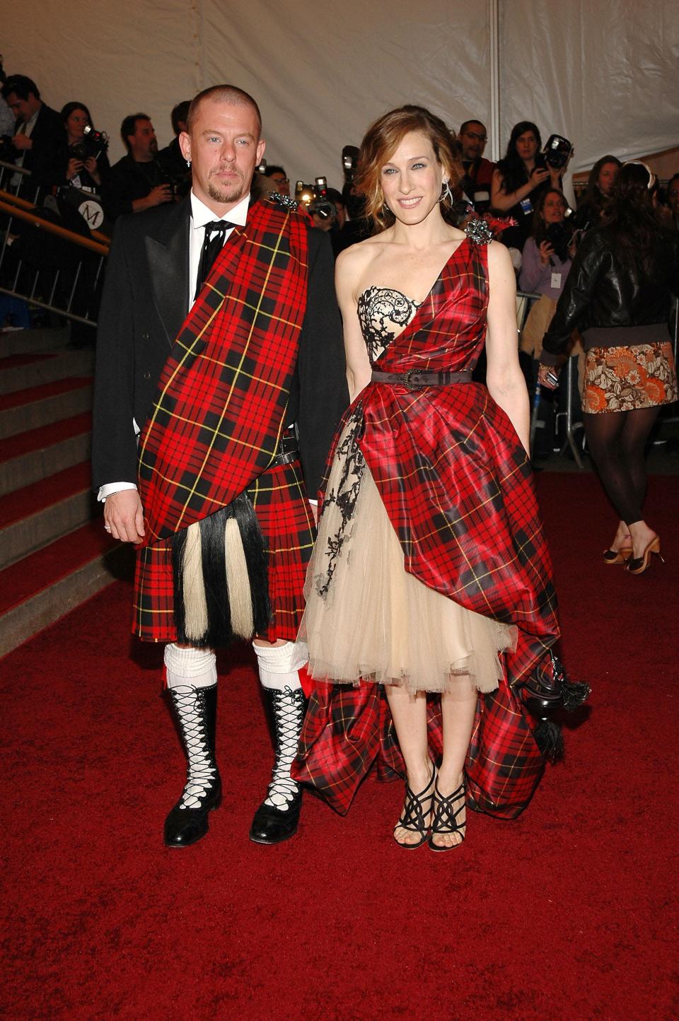 Alexander in a red plaid sash over his suit jacket and a red plaid kilt. He&#39;s standing side by side with Sarah Jessica in a nude tulle strapless dress with a lace bustier and red plaid sash forming one arm strap and being belted before flowing over the dress to the ground.