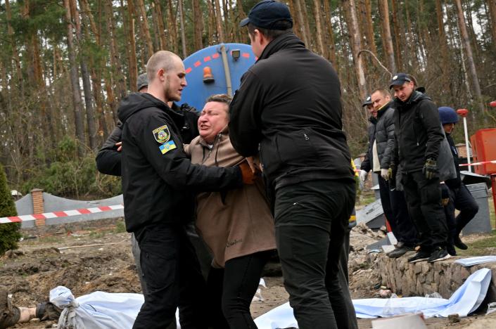 In a clearing in a wooded area, two men in their 20s try to hold up a Ukrainian woman, who wears an expression of extreme anguish and appears to be collapsing in shock.