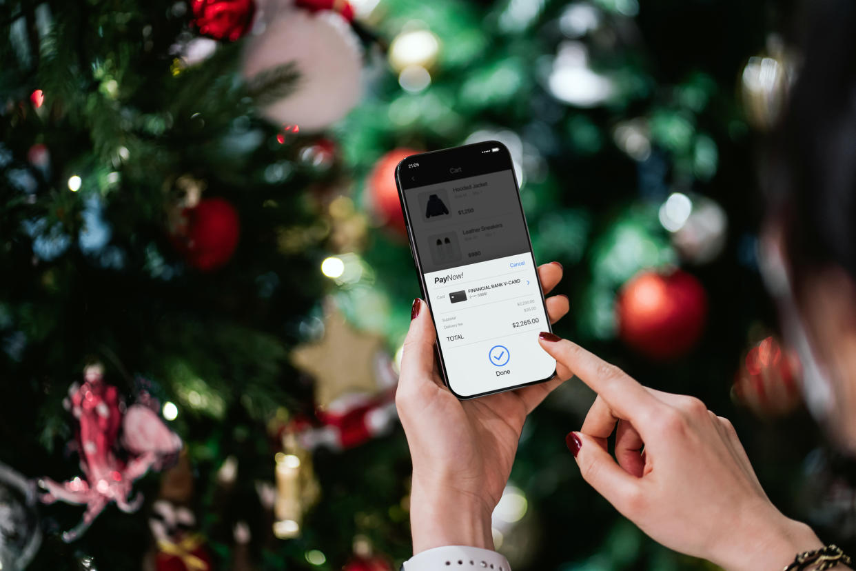  Over the shoulder view of woman's hand purchasing Christmas gift online with smartphone and making mobile payment with credit card against illuminated Christmas tree. . 