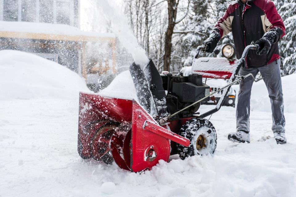 Man uses red and black snowblower with Jeep in parked in the backdrop. 