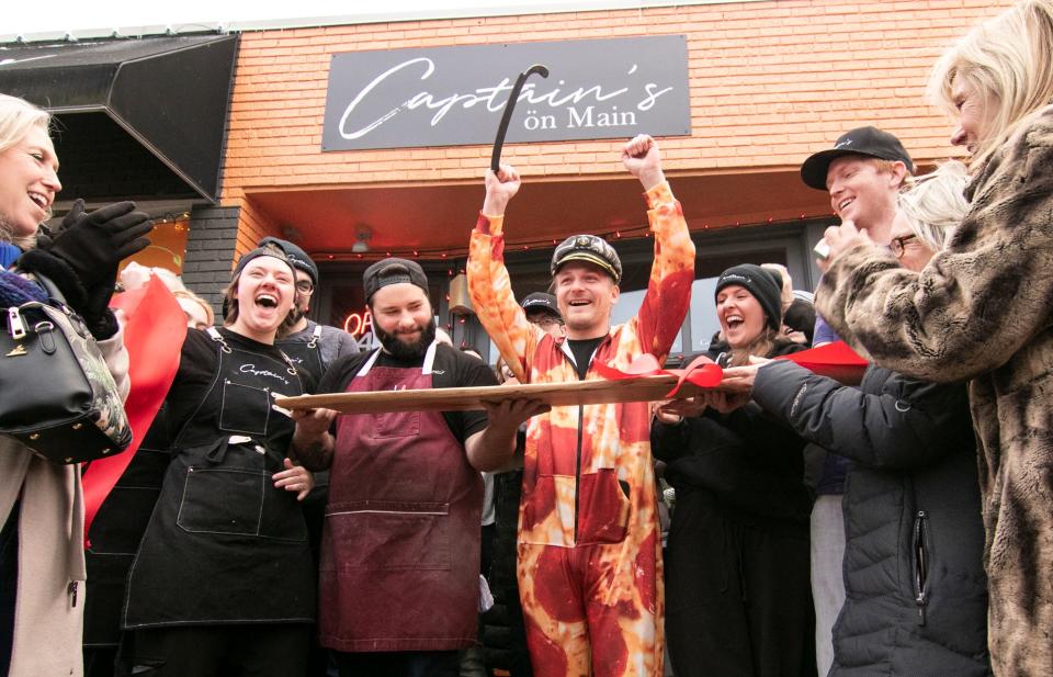 'Captain' Nick Mannisto (center) and his staff celebrate the ribbon 'slicing' in front of Captain's on Main in downtown Brighton Friday, Nov. 18, 2022. Mannisto used a pizza slicer to cut the ribbon against a commemorative cutting board that patrons and friends earlier signed with well wishes.