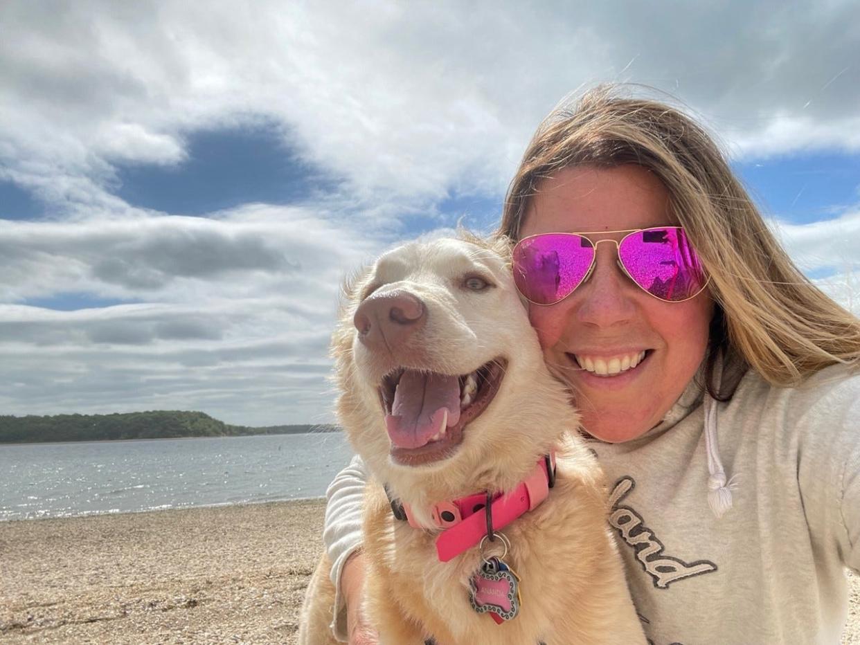 A white woman, Dina Pinelli, on the beach with sunglasses, with her dog, Ananda