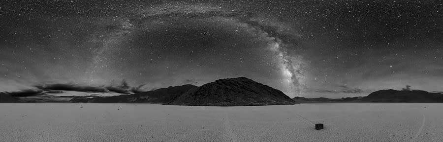 A black and white photo of the Milky Way taken in Death Valley over a playa with a rock seemingly moving across the ground with a track behind it. (Photo: D. Duriscoe / NPS)