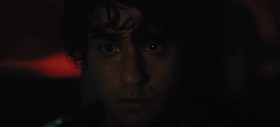 A teenage boy sits in a car, staring blackly in the distance