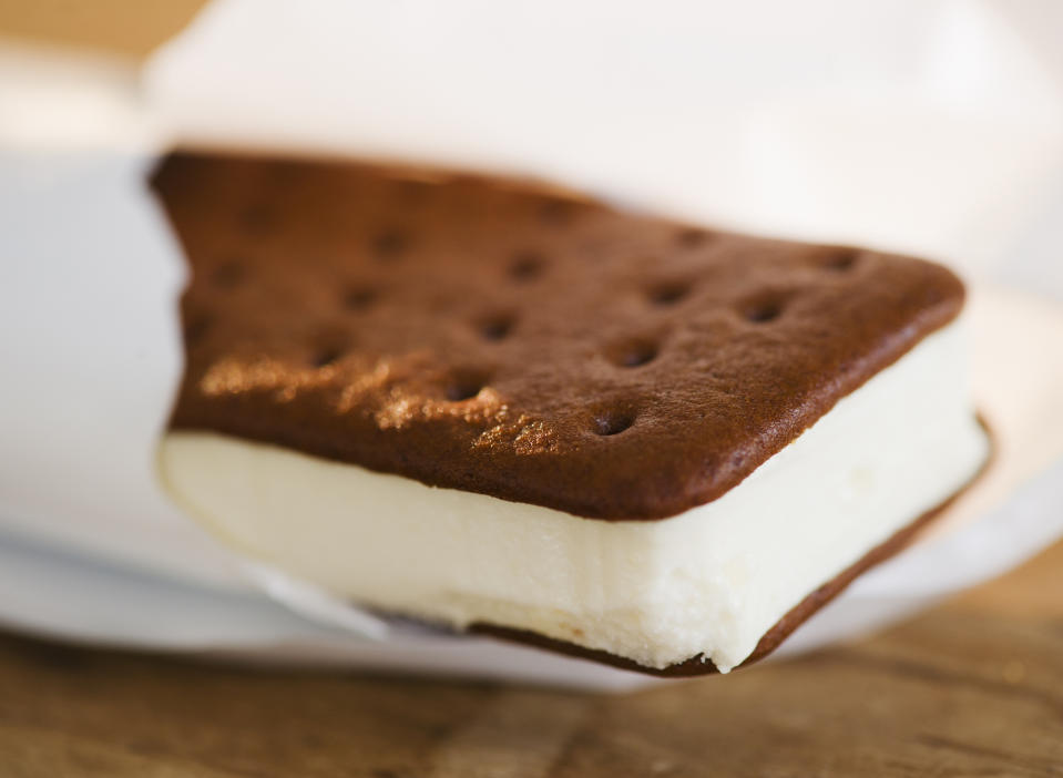 An ice cream sandwich partially unwrapped from its white paper wrapper, placed on a wooden surface